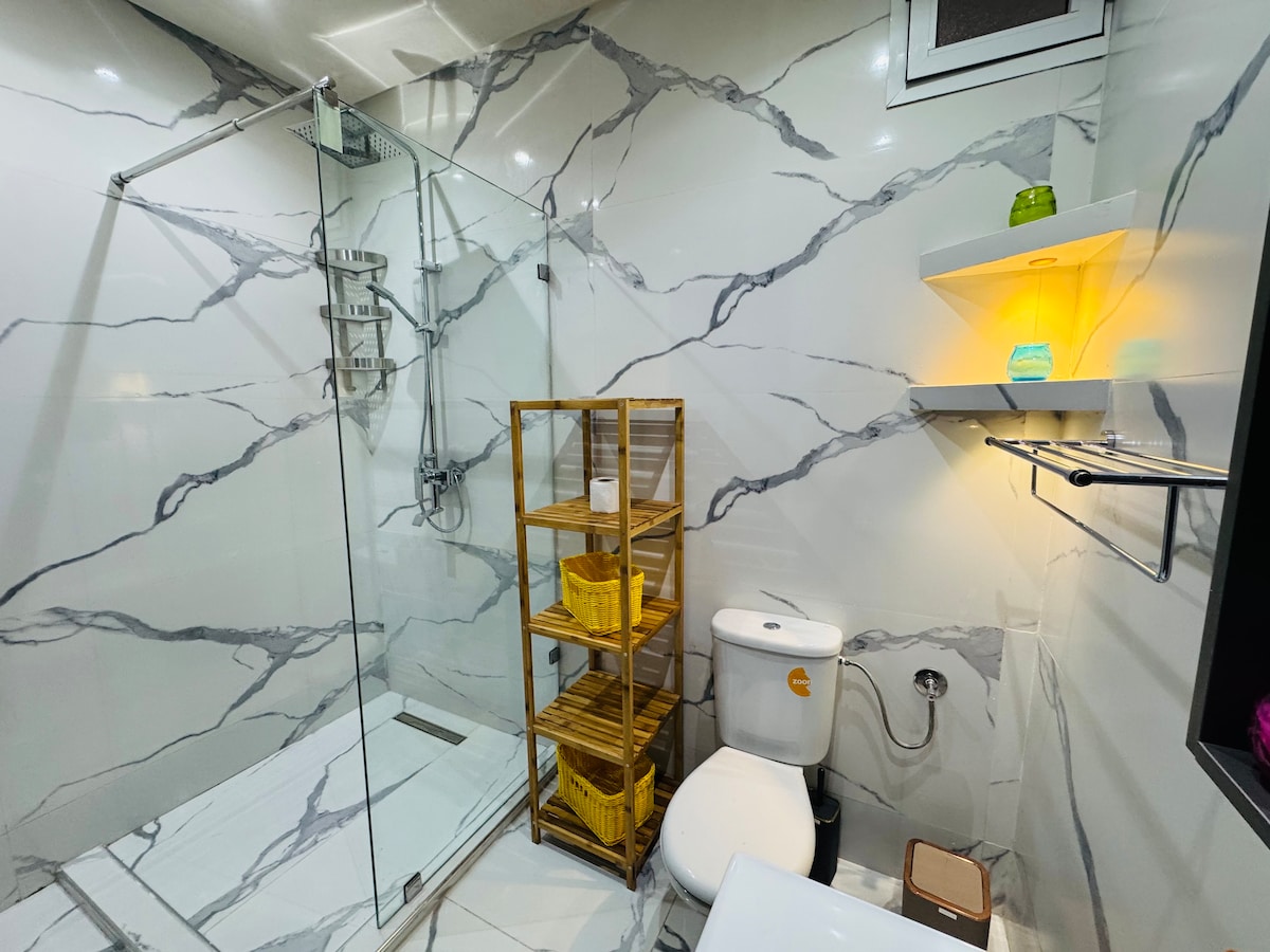 A modern bathroom is highlighted by sleek marble walls and a spacious walk-in shower featuring a glass enclosure. A wooden shelf unit is neatly arranged with storage baskets, while a bright light fixture adds warmth. A contemporary toilet is positioned for convenience.