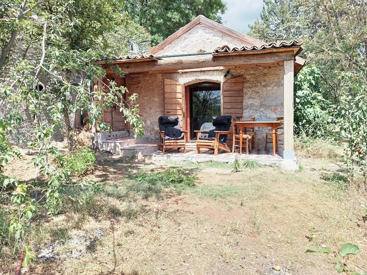The stone cottage features a wooden roof and an outdoor porch. Two comfortable armchairs are positioned beside a small wooden table, creating a cozy seating area. Surrounding greenery and trees offer a natural setting, with a clear view through the large glass door.