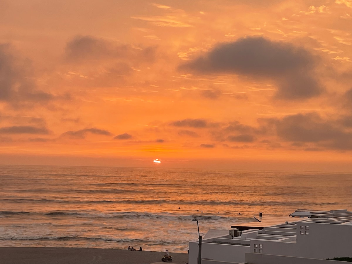 A serene ocean view captures a sunset illuminating the sky in shades of orange and yellow. Gentle waves reflect the warm colors, while silhouettes of a few people are visible along the shoreline. The tranquil scene conveys a sense of calm near the coastal area.