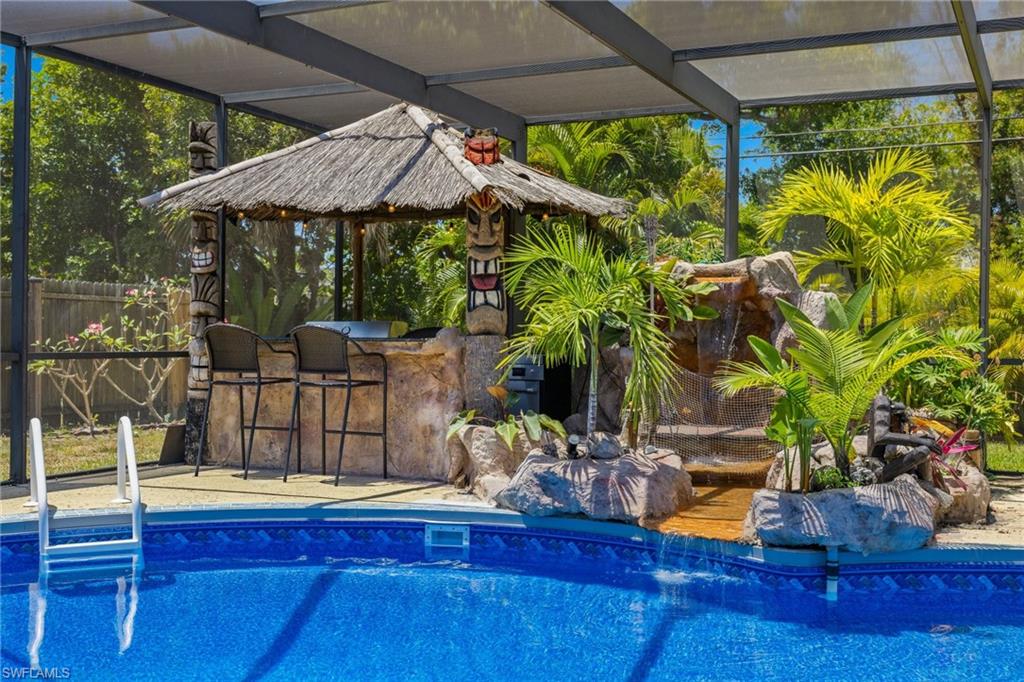 A custom tiki bar featuring a thatched roof and seating is positioned near a swimming pool. Tropical plants and a waterfall rock feature enhance the outdoor space, while the blue water of the pool sparkles in the sunlight.