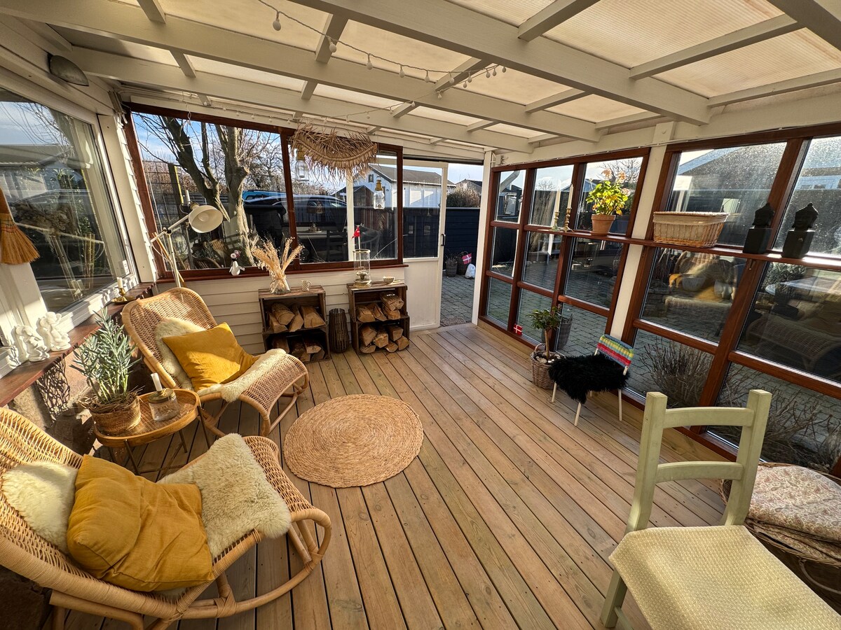 A large sunroom features wooden flooring and ample windows allowing natural light. Two rattan chairs and a small round rug are highlighted, with decorative items and logs displayed on a shelf. Potted plants add freshness to the inviting space.