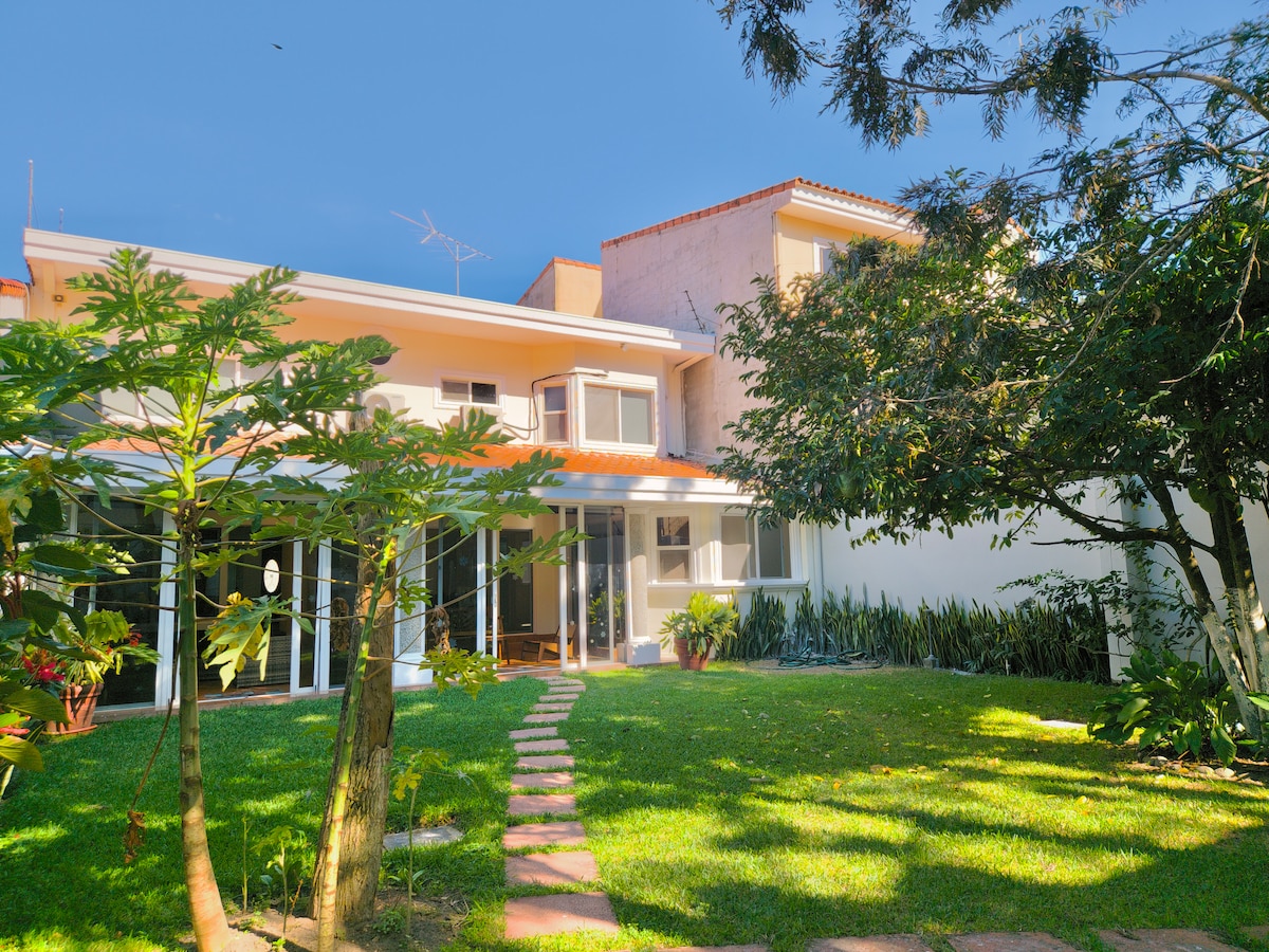The exterior of the house features a spacious garden with green grass and well-maintained trees. A stone pathway leads to the entrance, showcasing large windows that provide ample natural light to the living areas. The two-story structure is complemented by a bright blue sky.