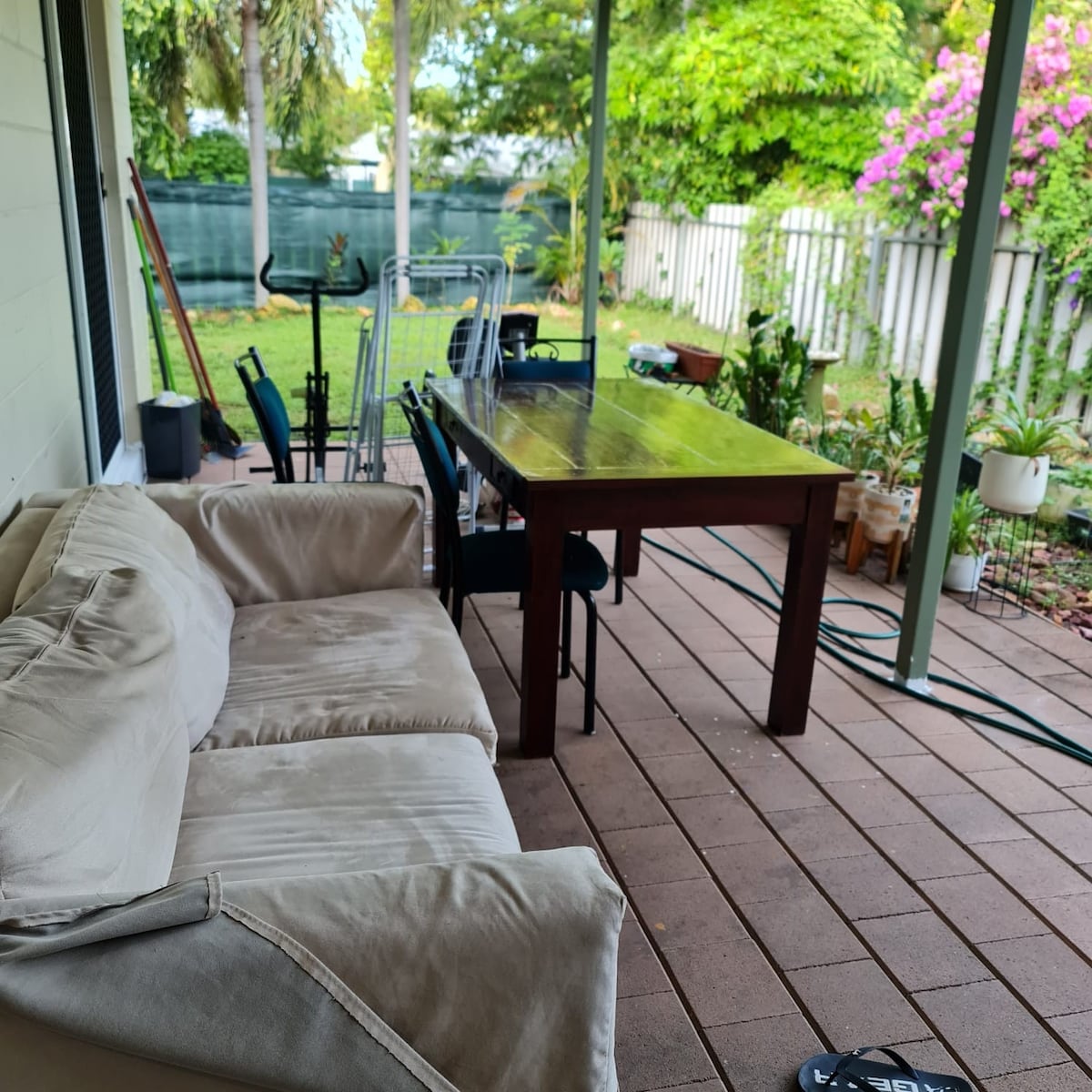 An outdoor patio area is depicted, featuring a wooden dining table surrounded by six chairs. A comfortable light-colored sofa is positioned along one side. Lush greenery and flowering plants add a natural ambiance, while a fenced garden area and storage items can be seen in the background.