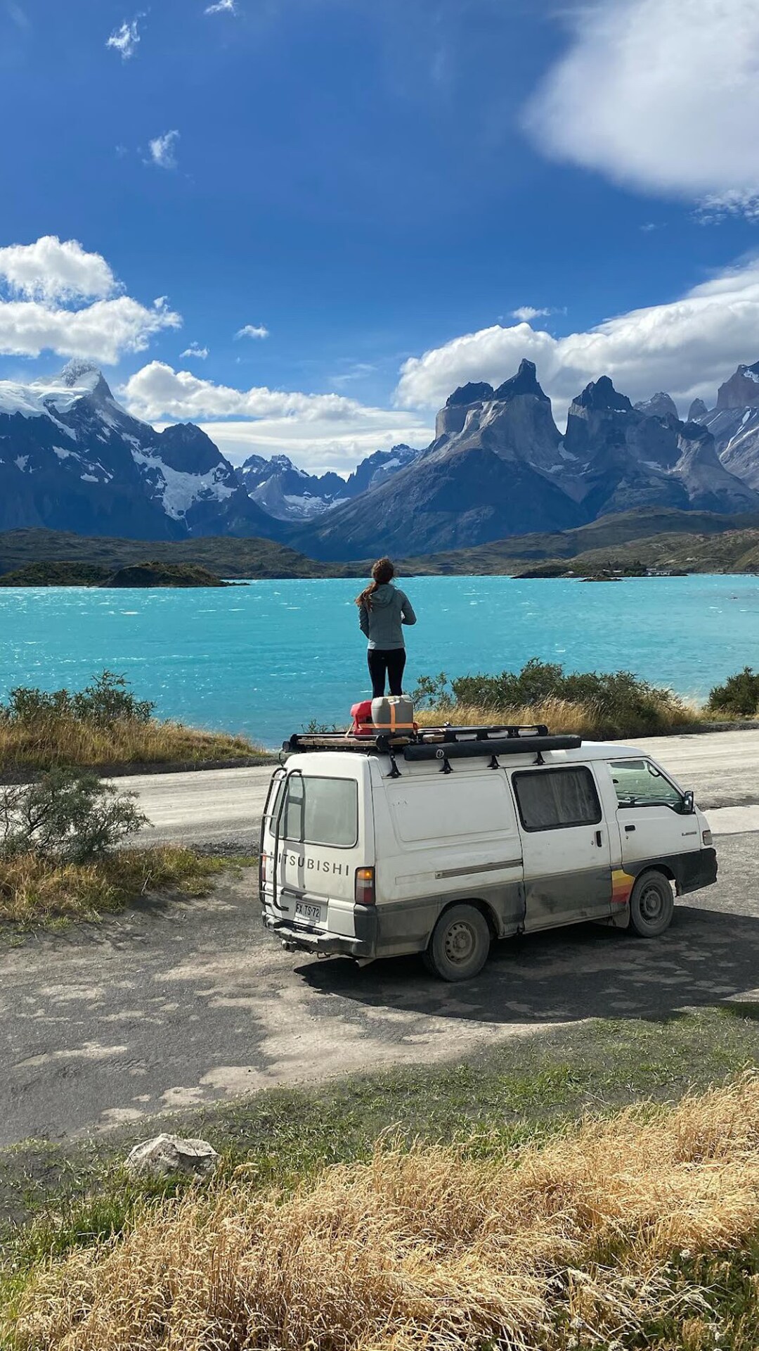 A compact campervan is parked by a turquoise lake, surrounded by dramatic mountains under a bright sky. A person stands atop the van, enjoying the panoramic view. Grassy terrain lines the foreground, enhancing the natural setting of the scene.