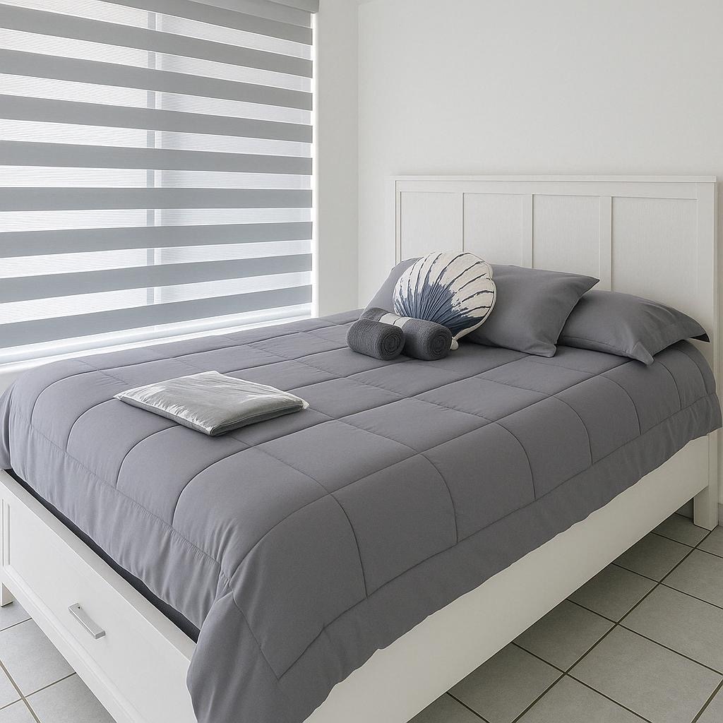 A cozy sleeping area features a spacious bed dressed in gray linens. Decorative pillows add a subtle touch, while light streams in through a modern striped window treatment. The floor showcases tiled flooring for a clean and minimalist aesthetic.