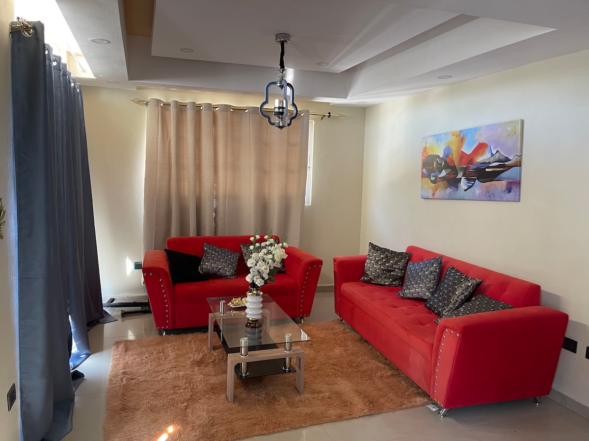 The living area features two red sofas adorned with decorative cushions, arranged around a glass coffee table. A plush rug covers the floor, while soft curtains frame the windows, allowing natural light to enhance the space. A colorful artwork hangs on the wall.