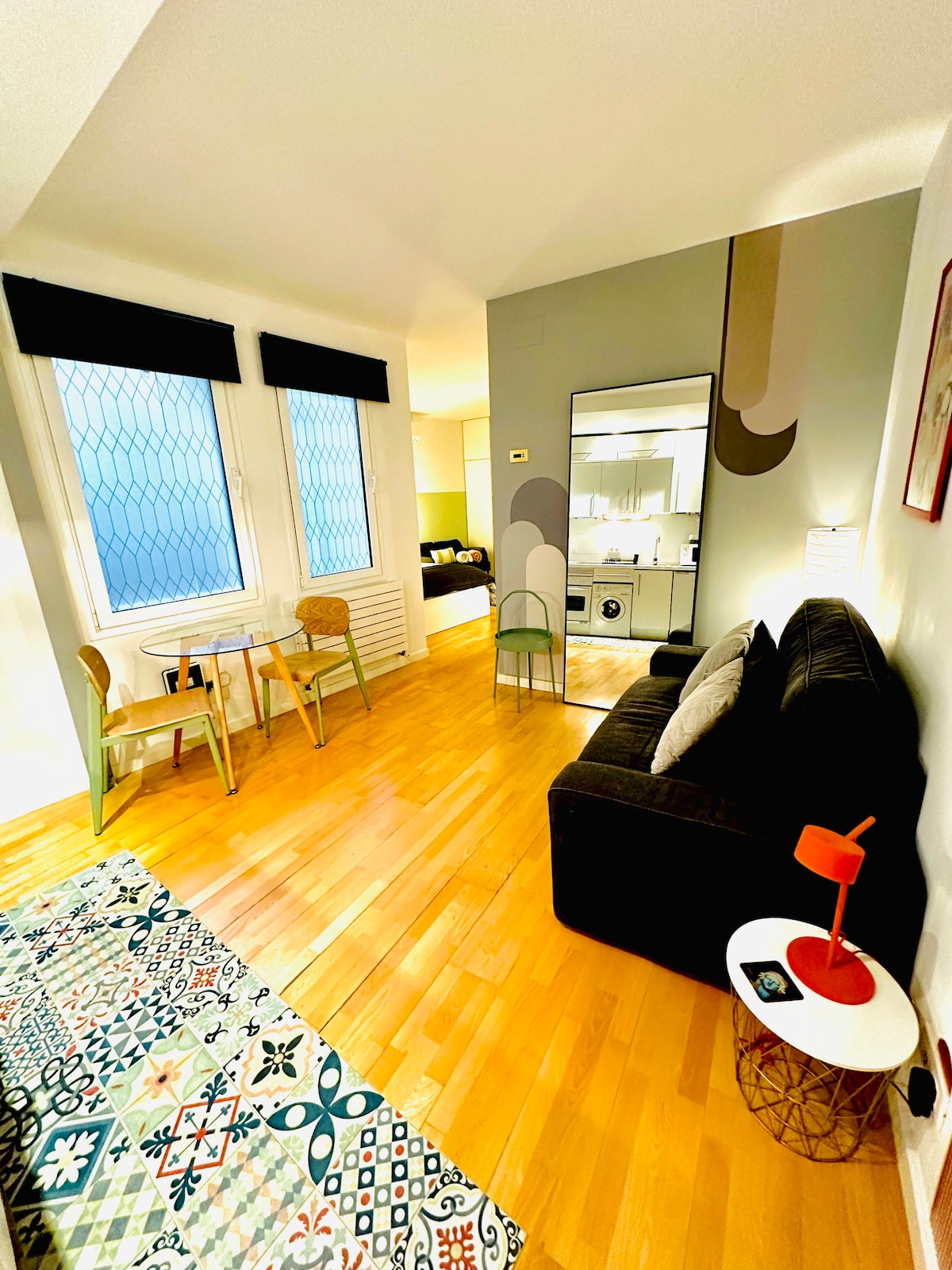 A modern living space features a dark sofa paired with minimalist chairs at a small dining table. Warm wooden floors are complemented by vibrant wall art. Natural light filters through patterned window coverings, and a mirror enhances the room's sense of openness.