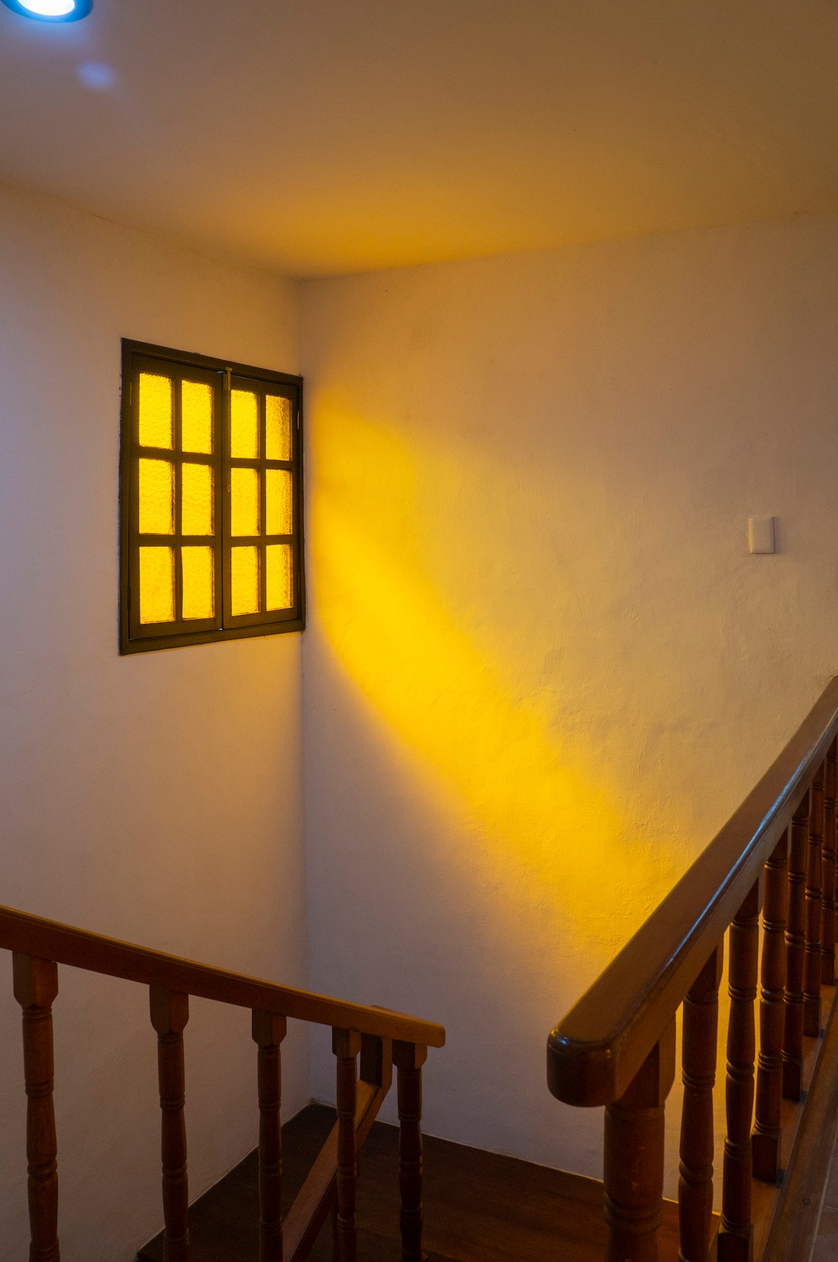 A staircase with wooden railings leads upward, illuminated by warm amber light filtering through a small window. The white walls enhance the brightness, creating a cozy, inviting atmosphere that guides guests to the upper levels.