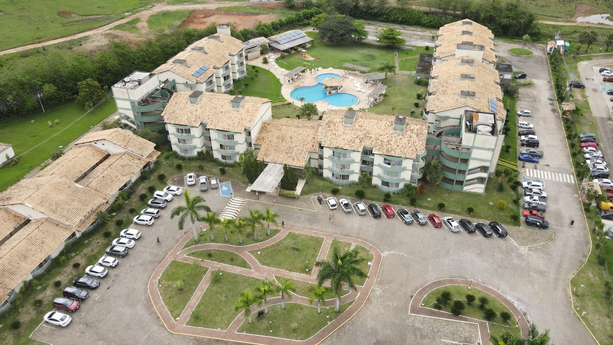 An aerial view captures the layout of the Village das Águas condominium, featuring multiple buildings with green accents. A central pool is visible, surrounded by landscaped areas and a parking lot filled with vehicles. Lush greenery enhances the setting.