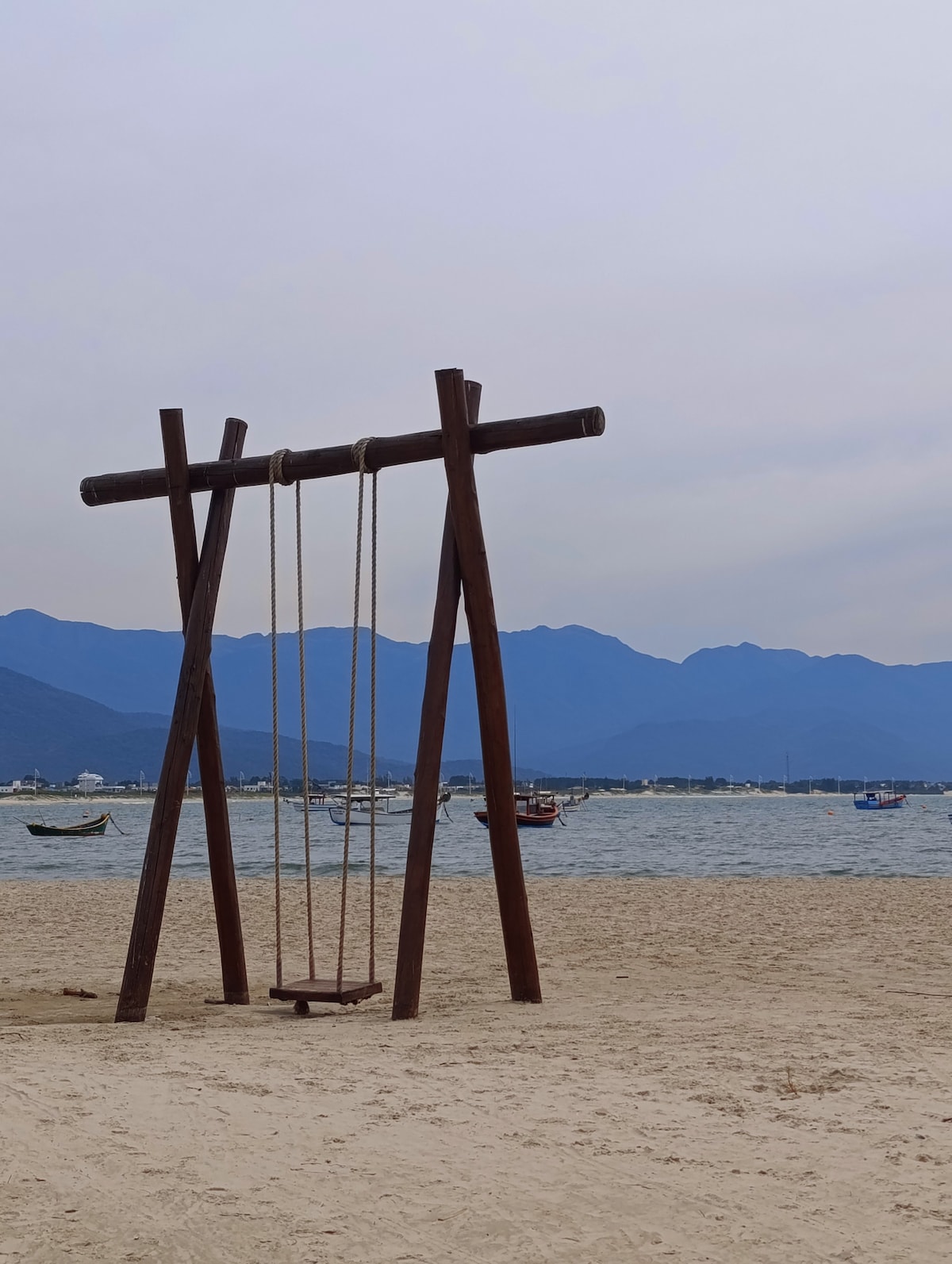 A wooden swing set stands on a sandy beach, with mountains in the background. The calm water reflects the soft hues of the sky. Several boats can be seen floating in the distance, framed by a serene coastal atmosphere.