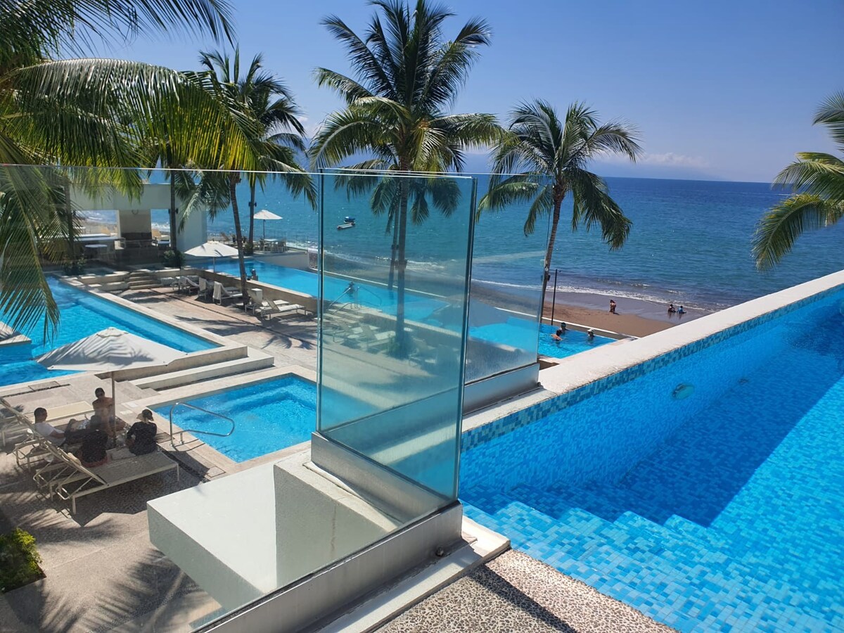 Multiple inviting pools are visible, surrounded by palm trees and lounge chairs. The shimmering blue water reflects sunlight, while the beach and ocean lie just beyond. A clear view of the horizon showcases the serene beachfront environment.