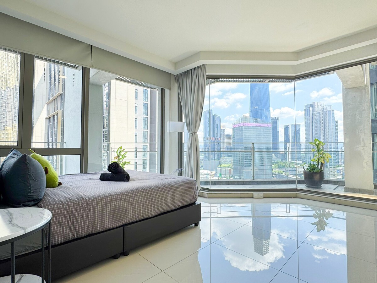 A bright bedroom features a spacious bed with neutral bedding and decorative pillows. Floor-to-ceiling windows offer a panoramic view of the city skyline and allow natural light to fill the room. A potted plant adds a touch of greenery to the contemporary decor.