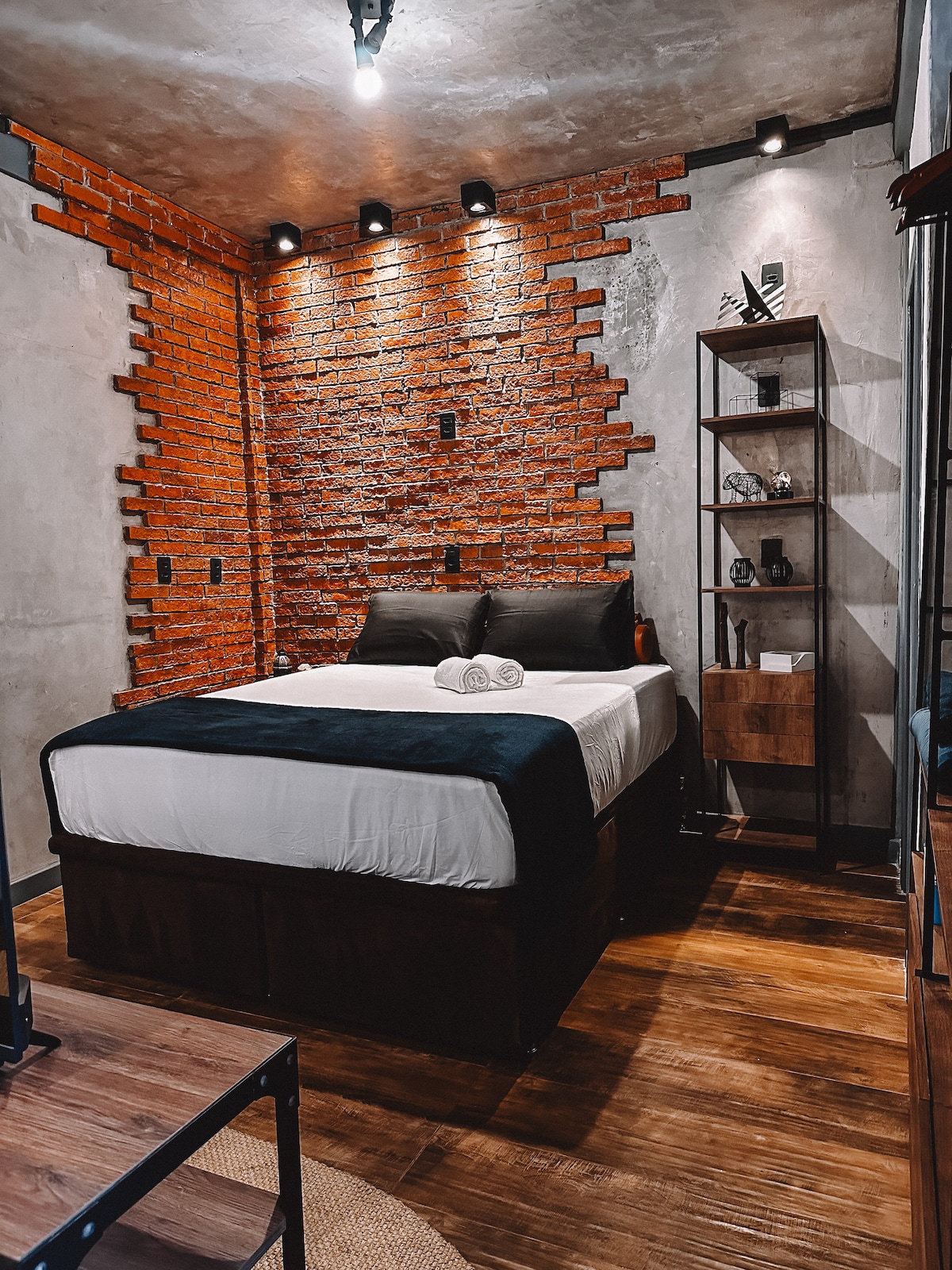 A modern bedroom features a spacious bed with dark linens, flanked by decorative pillows. Exposed brick walls create a bold accent, while warm lighting is mounted above. A wooden shelf displays decorative items, and a wooden tabletop adds to the contemporary design.