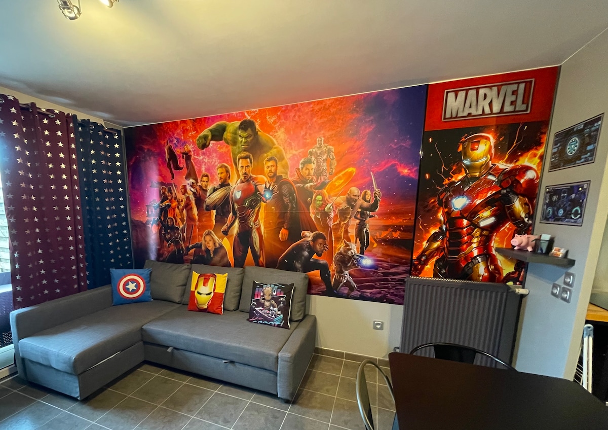 A living area features a spacious grey sofa positioned next to a vibrant Marvel-themed wall mural, showcasing superheroes in action. Decorative cushions add colour and character. Soft lighting highlights the modern design, while a small dining table is visible in the background.