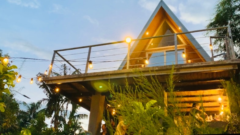 The triangular structure features a wooden deck adorned with string lights along the railing. Large windows create a bright atmosphere, allowing glimpses of the interior. Surrounding tropical plants add greenery, establishing a connection to the garden below.
