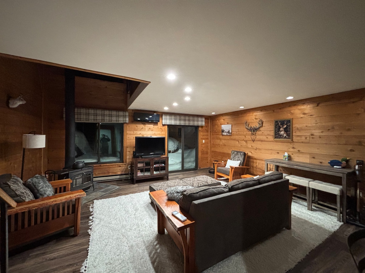 A spacious living area is highlighted by wooden wall paneling and large windows with plaid valances. A comfortable sofa and chairs surround a coffee table, creating an inviting seating arrangement. A TV is mounted on the wall, and a dining table is present in the background.