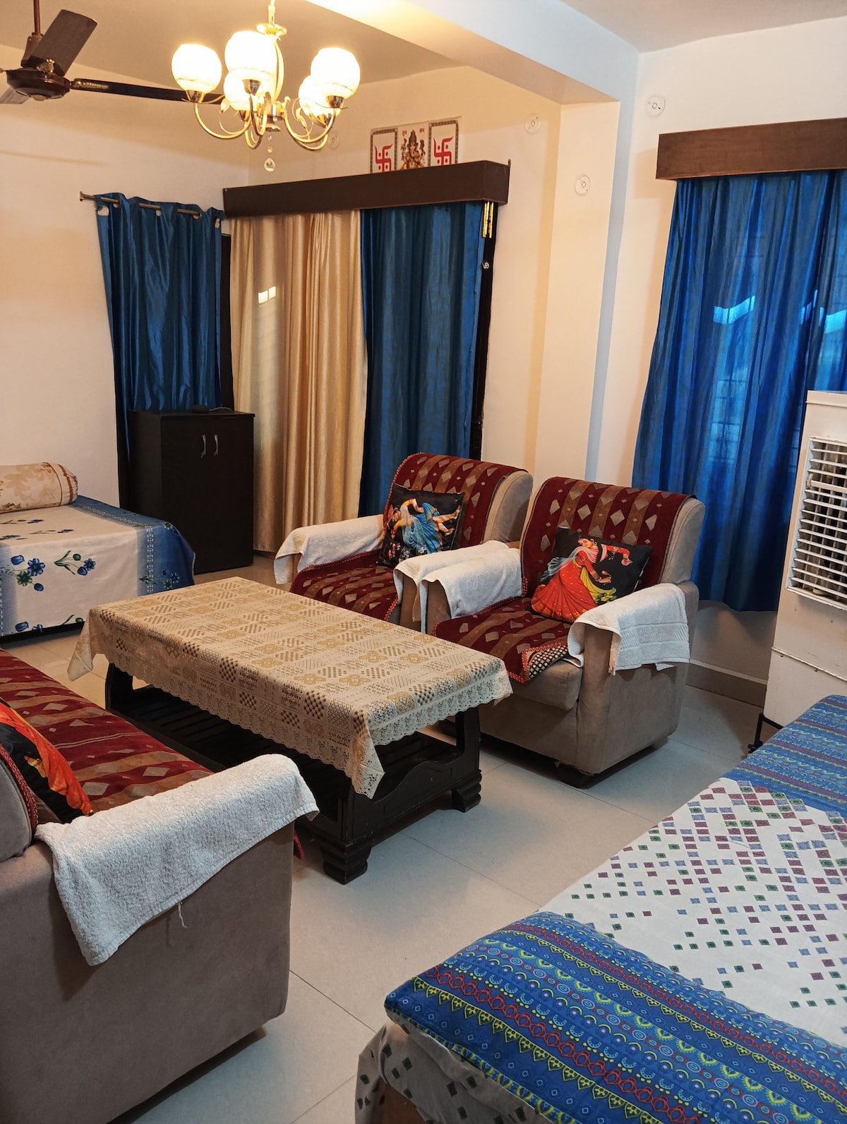 A living area features two plush armchairs, with decorative throws draped over them. A wooden coffee table sits at the center, surrounded by soft lighting from a ceiling fixture. Light blue curtains frame the windows, complementing the muted interior tones.