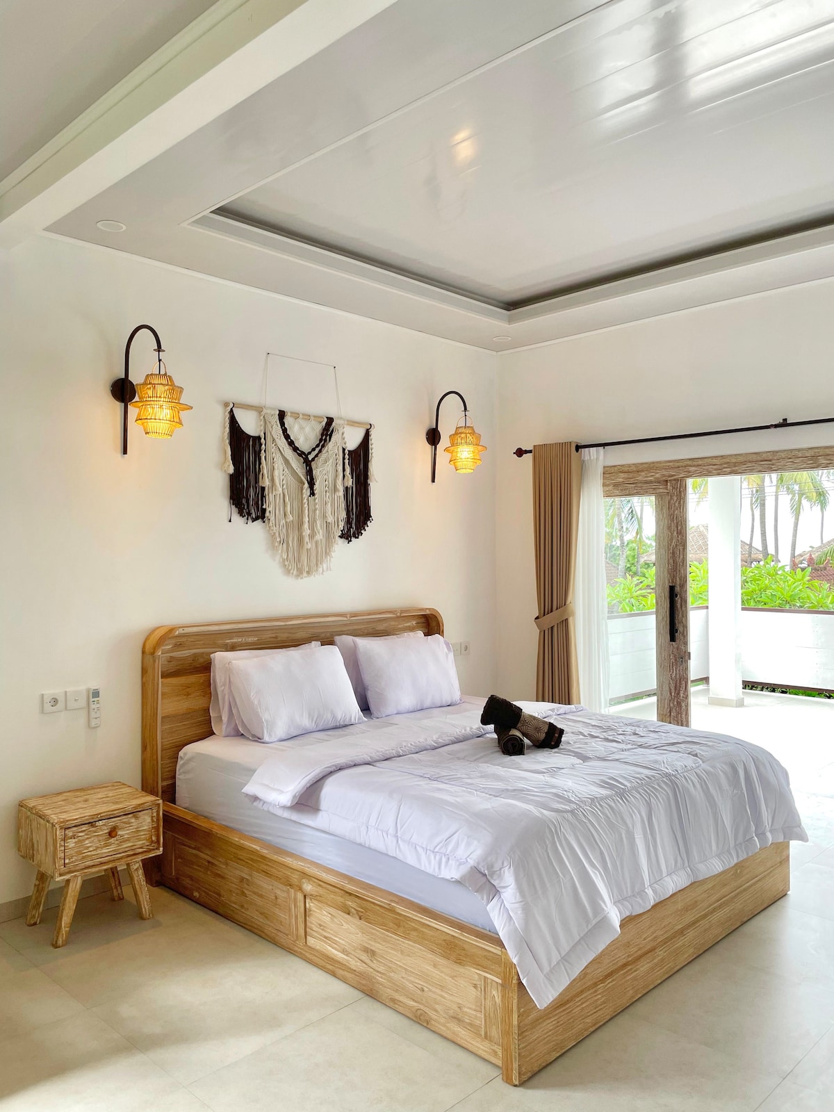 A spacious bedroom features a wooden bed with neatly arranged linens. Soft lighting fixtures are mounted on the walls, and a decorative wall hanging adds visual interest. Large glass doors provide a view of the outdoor area, allowing natural light to fill the room.