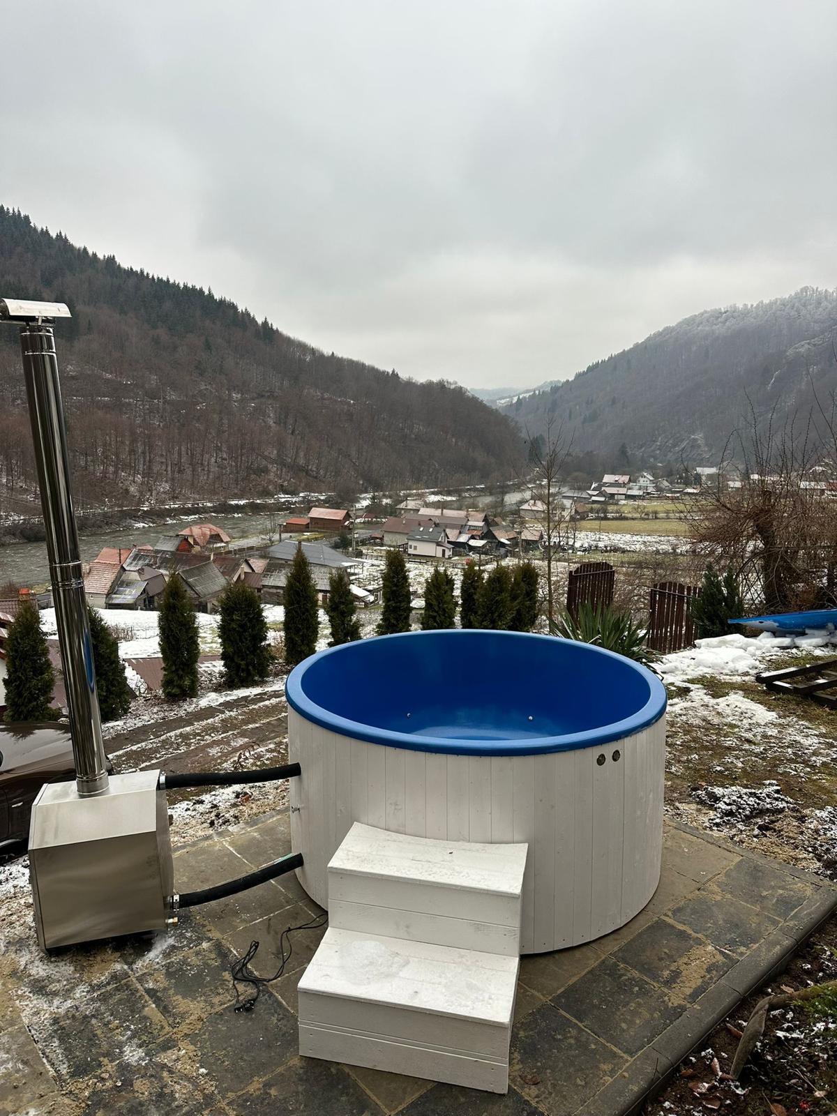 An outdoor hot tub with a blue rim is situated on a stone patio, overlooking a valley covered in a light dusting of snow. Steps lead up to the hot tub, while evergreen trees frame the scenic view of the distant hills and picturesque village below.