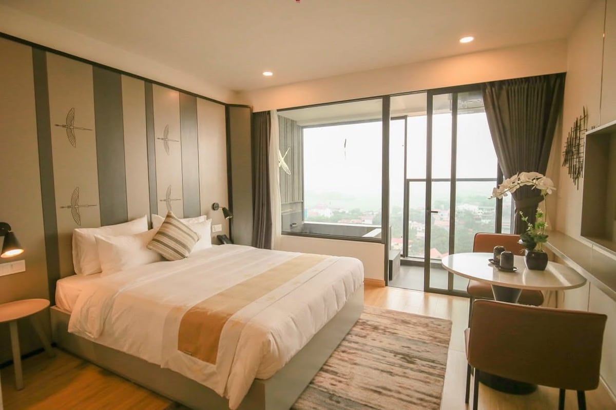 A well-appointed bedroom features a spacious bed with neutral-toned bedding. A sitting area by the window overlooks distant landscapes. Natural light floods the room through floor-to-ceiling glass doors, complemented by elegant decor and a small dining table with two chairs.