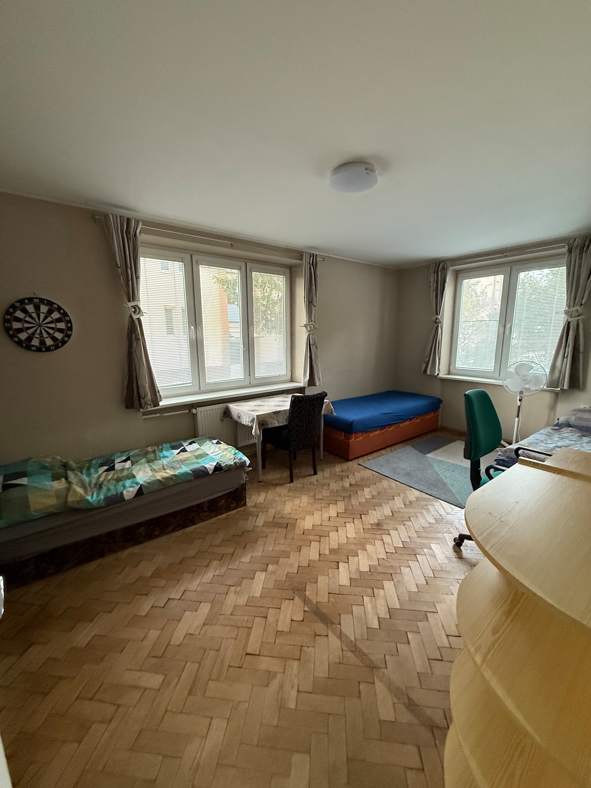 A well-lit room features two beds, one of which is positioned against a wall. A small table with a chair is situated near the window, allowing natural light to fill the space. A wardrobe is present, along with a rug on the floor.