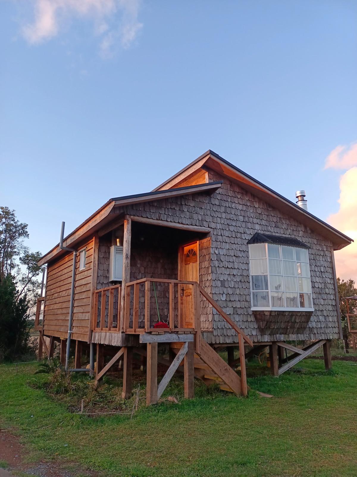 A rustic cabin constructed primarily of wood features a shingled exterior. The entrance is accessible via wooden steps, and a large window allows natural light to illuminate the interior. Surrounding greenery provides a serene atmosphere.