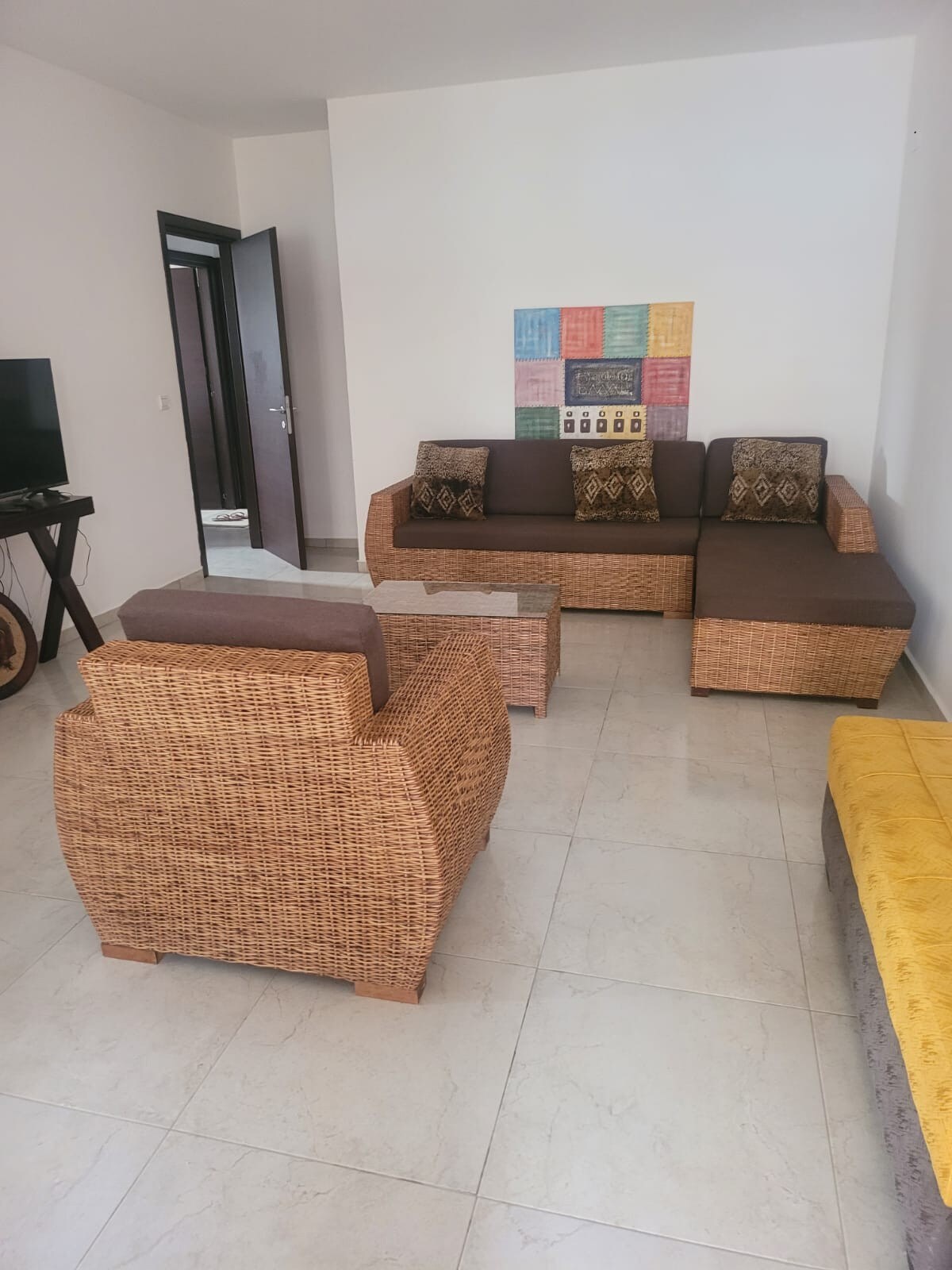 A spacious living area displays a woven rattan sofa and armchairs, complemented by decorative cushions. A rectangular coffee table sits in the center, while a colorful wall art piece adds interest. The light-colored tiled floor enhances the room's bright and airy feel.