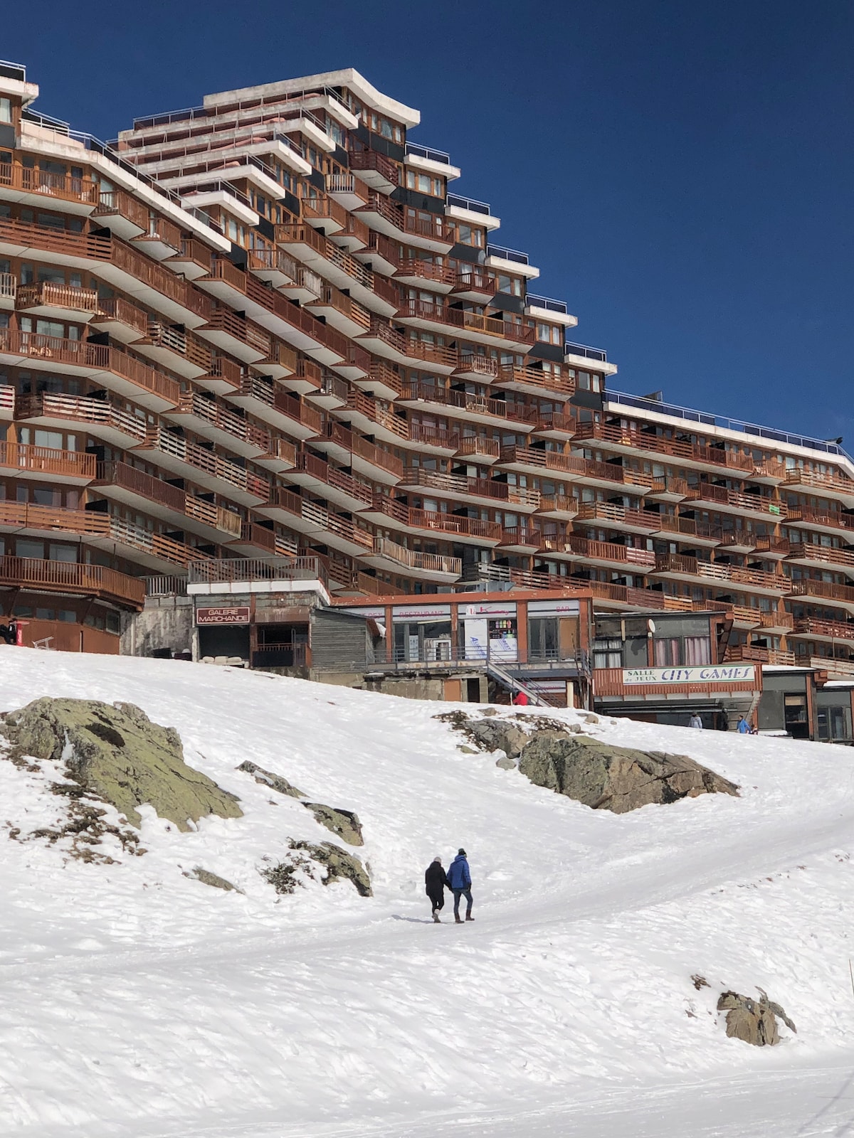 A multi-story building featuring numerous balconies is visible against a clear blue sky. A snow-covered pathway leads towards the entrance, while two individuals walk along the path, highlighting the wintry setting. The surrounding landscape is blanketed in snow, creating a serene atmosphere.