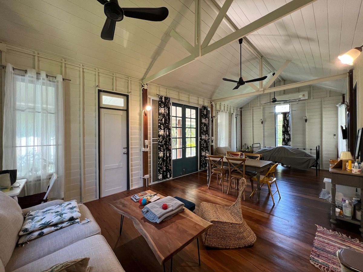 An open living area features wooden flooring and high ceilings with exposed beams. A light-colored sofa and a rustic coffee table are positioned adjacent to a dining table surrounded by chairs. Two ceiling fans are visible, providing a cool ambiance.