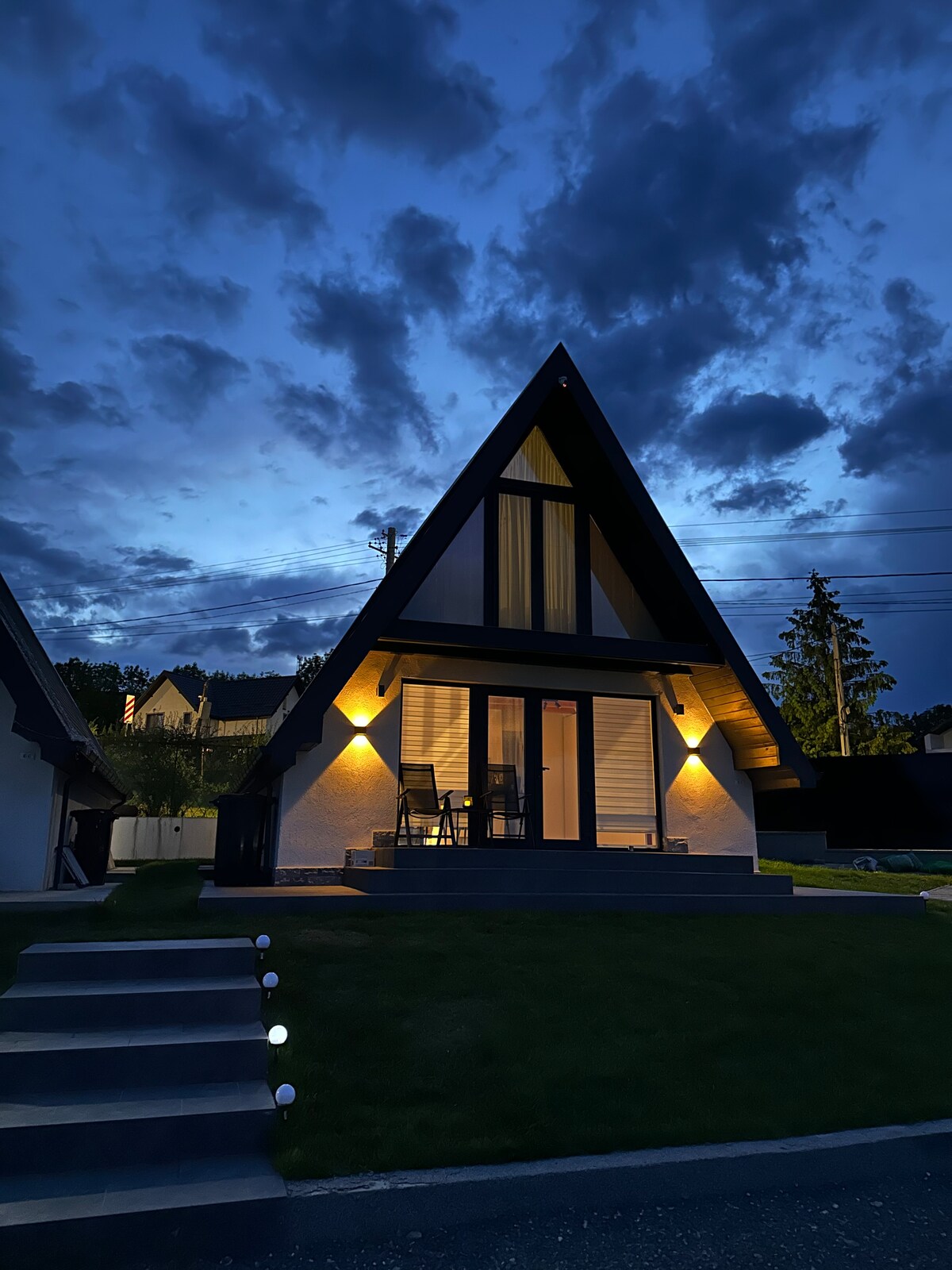 An A-frame cabin is shown under a twilight sky, featuring a distinctive triangular roof and large glass doors. Soft exterior lighting highlights the entrance, while a small staircase leads to a green lawn. The surrounding trees are partially visible in the background.