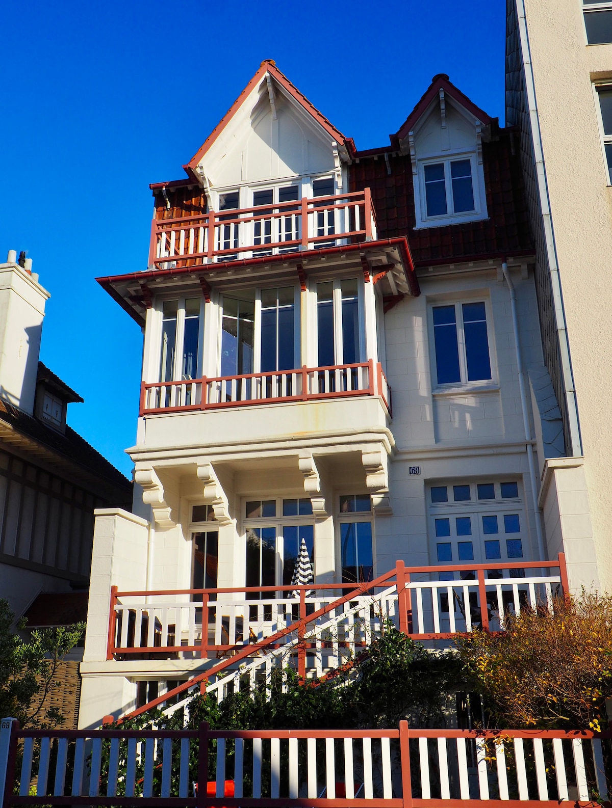 A three-story villa is presented with a distinctive architectural style. Large windows allow natural light to fill the interior. A red-tiled roof complements the white exterior. A staircase leads to the entrance, framed by a white picket fence and surrounding greenery.
