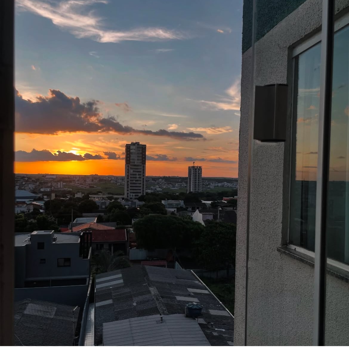 A sunset view is captured from a balcony, displaying vibrant hues of orange and yellow across the sky. A cityscape in the foreground includes a mix of residential buildings and rooftops, while distant buildings stand against the horizon.