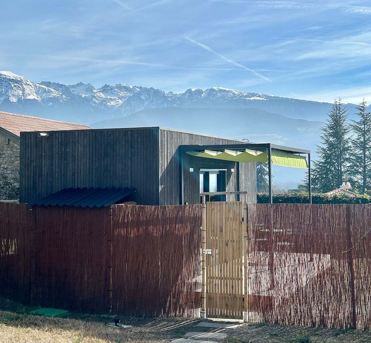 A modern tiny house is surrounded by a wooden fence, featuring a secure gate. Snow-capped mountains are visible in the background, complemented by a clear blue sky. The house's exterior showcases large windows and a covered patio for outdoor enjoyment.