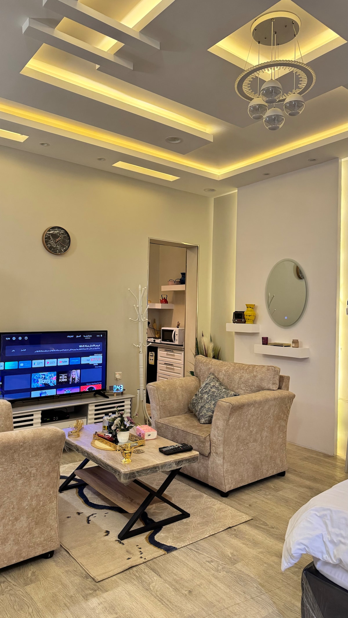 A modern living area features two plush chairs next to a low coffee table, with a sleek TV mounted on the wall. A round mirror and minimalist shelf enhance the space, while soft lighting from the ceiling creates a welcoming atmosphere.
