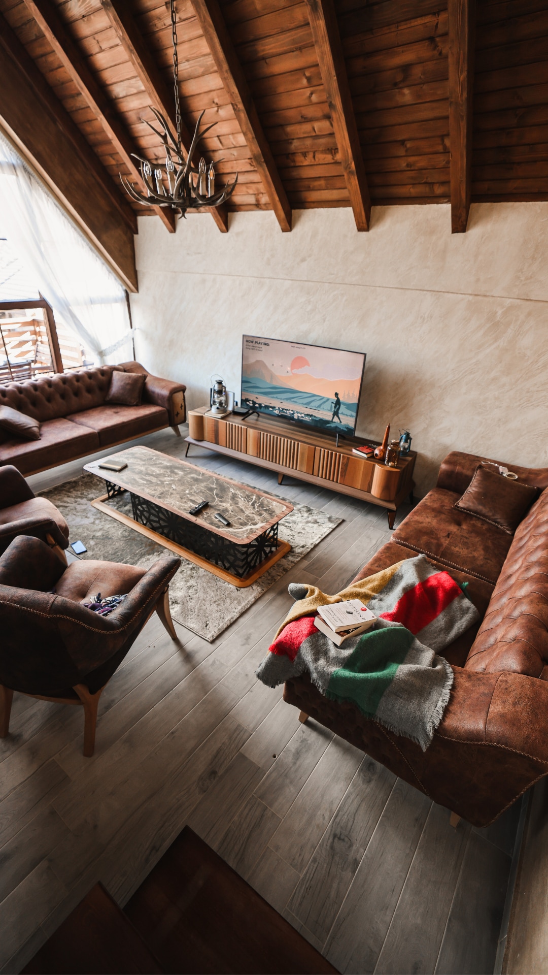 A spacious living area features high wooden ceilings and ample natural light from large windows. Two leather sofas and individual chairs provide seating around a central coffee table. A warm throw and books are casually arranged on one sofa, while a television is mounted on the wall.