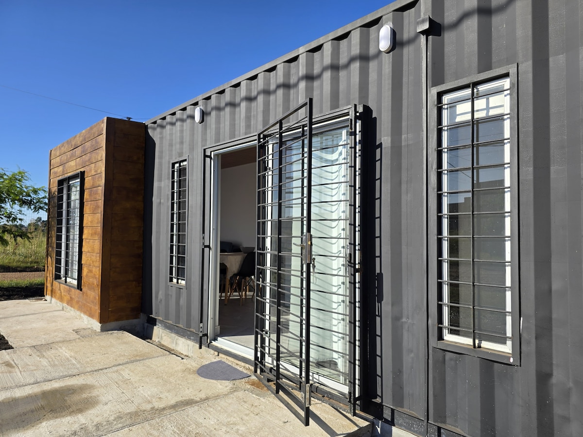 The exterior features a modern design with a combination of dark containers and wooden paneling. Double glass doors open to a welcoming interior, flanked by several windows with grid patterns allowing natural light. A smooth concrete pathway leads to the entrance.
