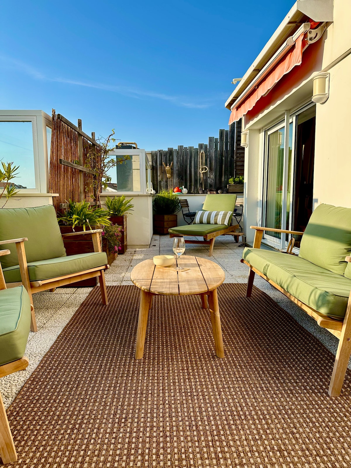 An inviting terrace features comfortable seating arranged around a low wooden table. Lush greenery in pots adds a natural element, complemented by a clear blue sky above. A cozy ambiance is enhanced by sunlight illuminating the space. Perfect for outdoor relaxation.