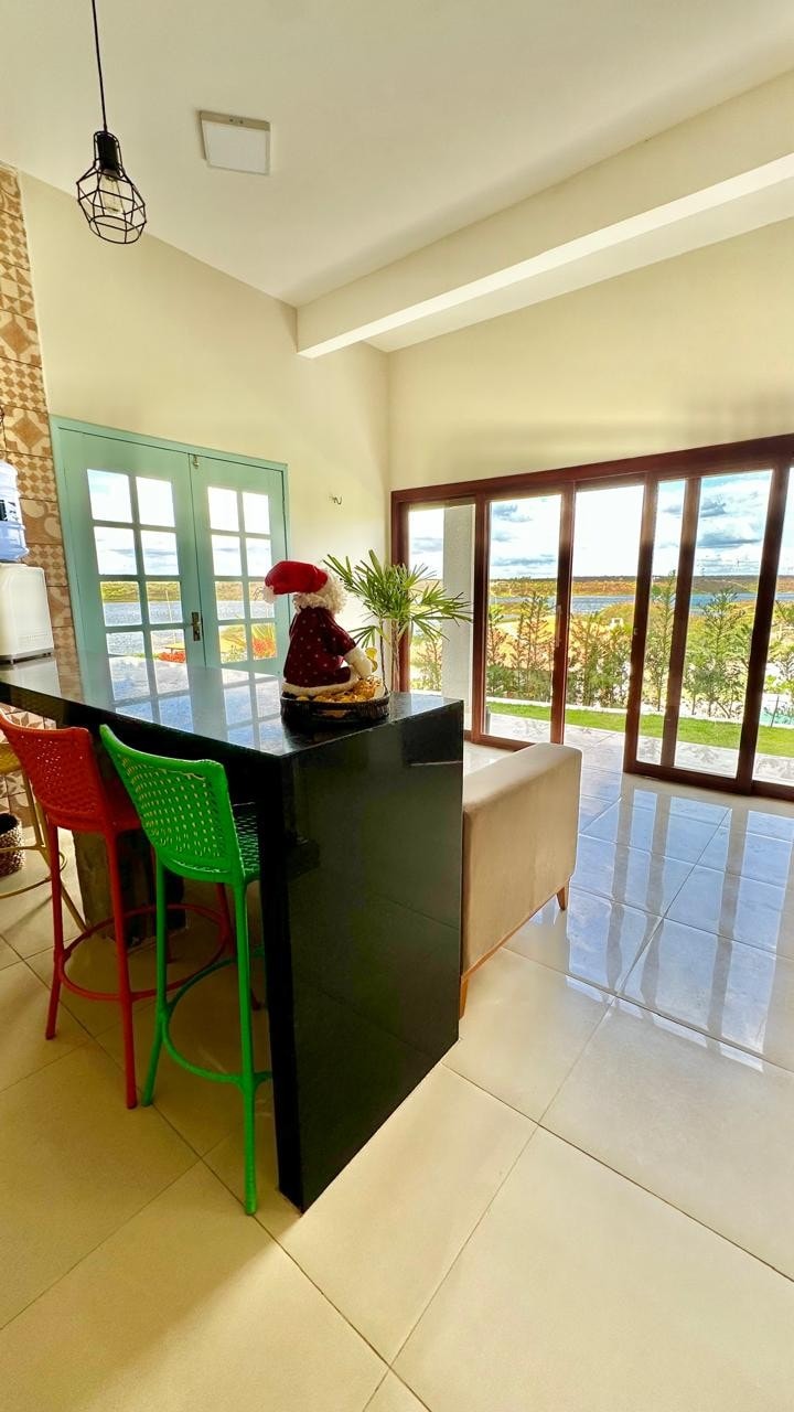 A kitchen area is depicted, featuring a black countertop and colorful bar stools in green and red. Large glass doors open to a scenic view, inviting natural light into the space. The modern decor includes potted plants, creating a fresh and bright atmosphere.