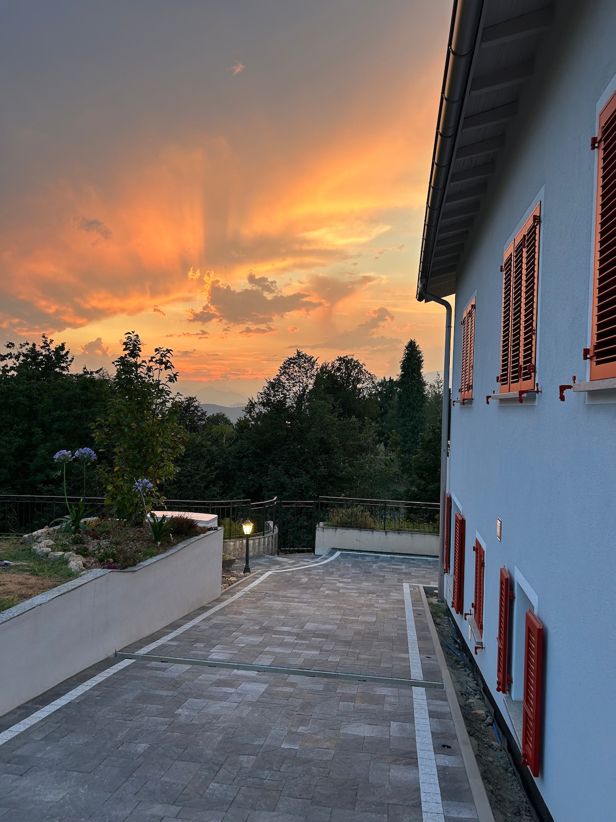 The exterior of the house is shown against a vibrant sunset, highlighting the soft hues of the sky. The building features striking red shutters and is surrounded by greenery. A paved pathway leads to the entrance, providing a welcoming view of the surrounding landscape.