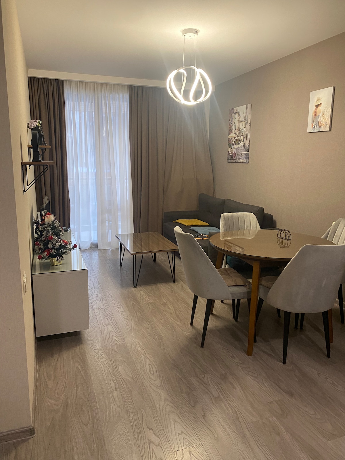 A modern living area with a light fixture overhead features a gray sofa and a round dining table surrounded by six chairs. Natural light enters through the sheer curtains, enhancing the neutral color palette of the space. Decorative elements include floral arrangements and artwork on the walls.
