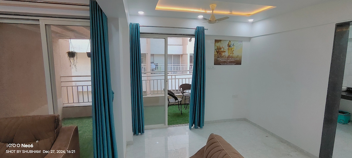 A well-lit living area features large glass doors that open to a balcony, offering natural light. Blue curtains frame the windows, and a comfortable sofa is positioned towards the center. A potted plant is visible outside, with a chair set near the balcony.