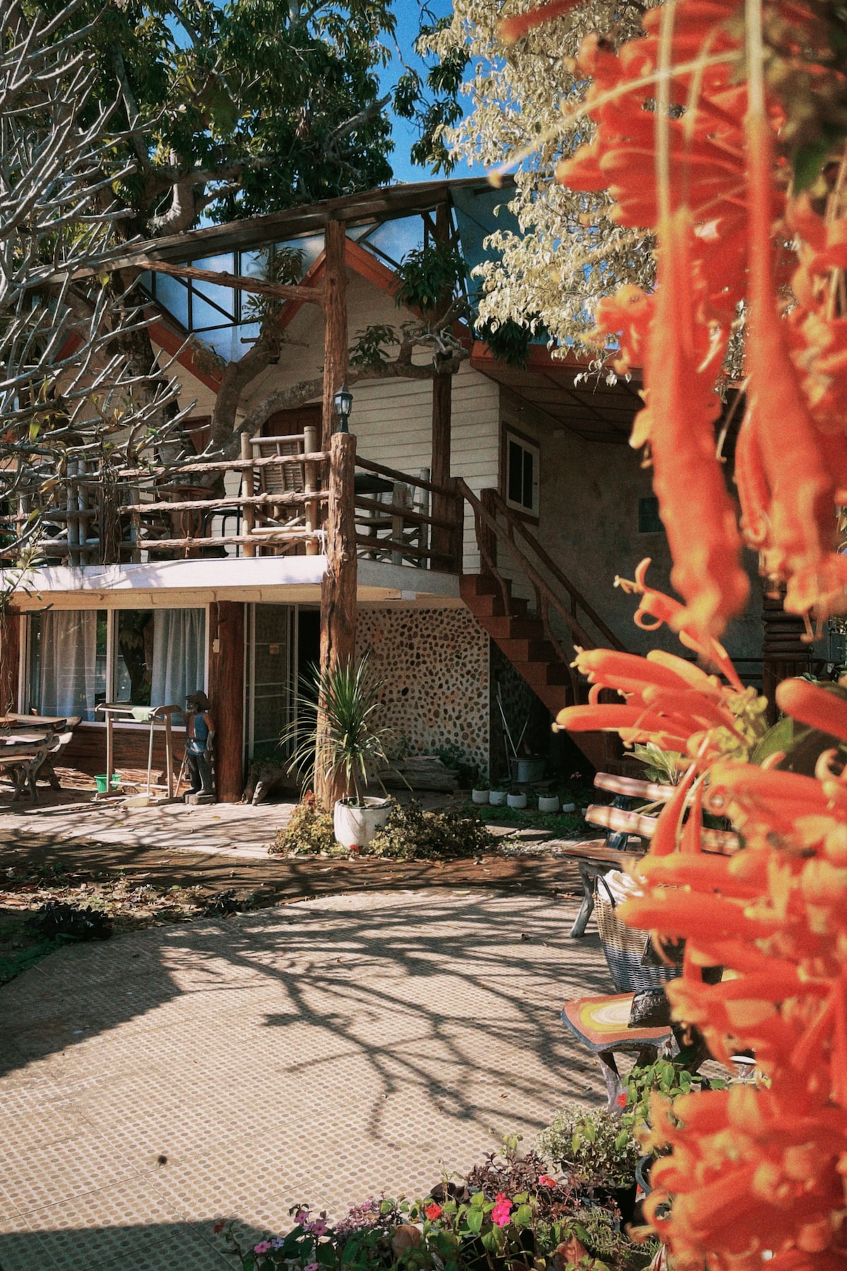 A unique, two-story structure is framed by vibrant orange flowers and greenery. The outdoor space features a tiled patio, wooden railings, and steps leading to a second-floor deck. Various plants and decorative elements enhance the serene environment, inviting relaxation.