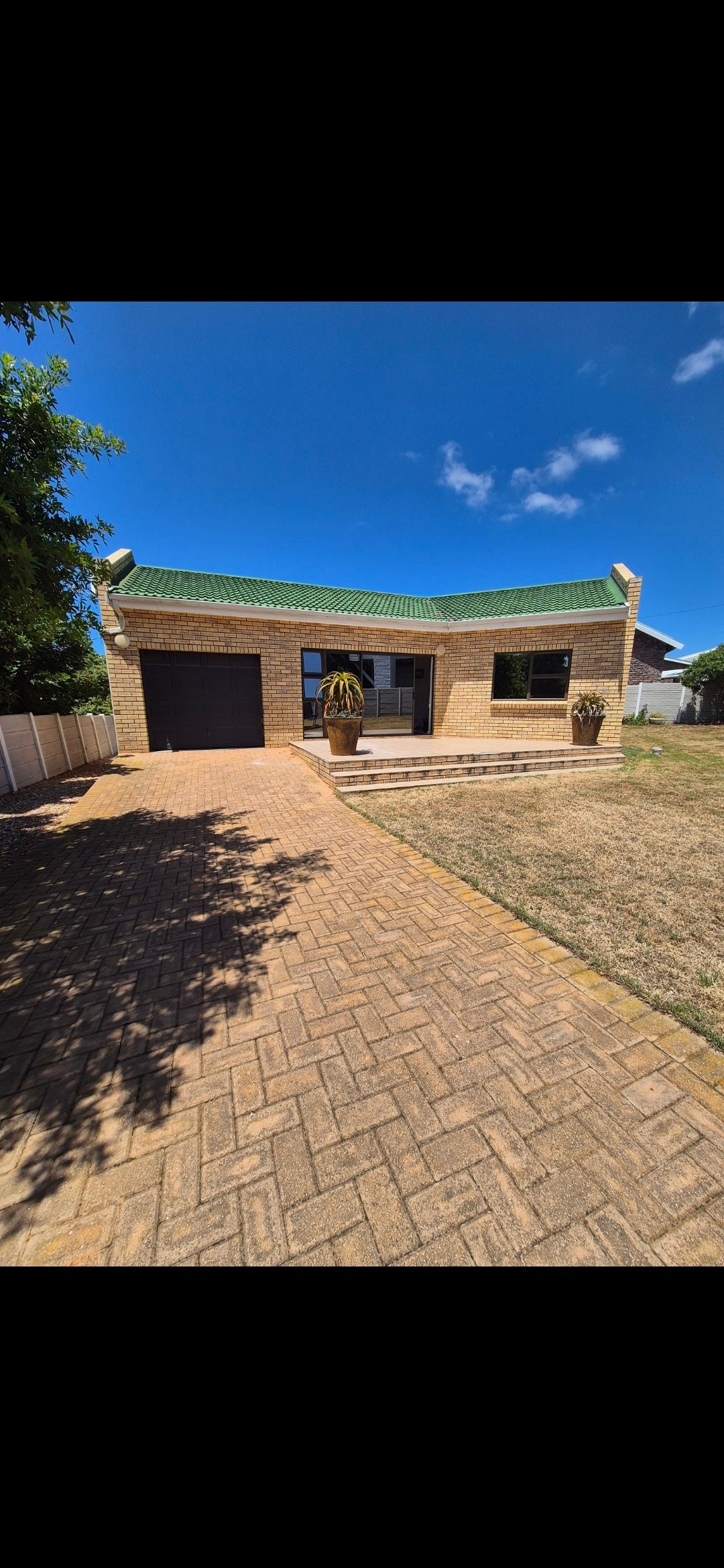 A single-story cottage is visible with a brick pathway leading to the entrance. The exterior features a green roof and tan bricks, complemented by potted plants flanking the front door. A clear blue sky serves as the backdrop, enhancing the cottage's inviting presence.