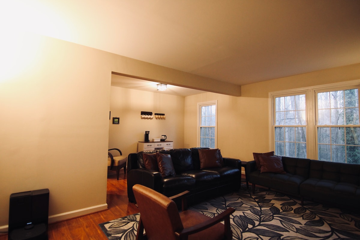 A spacious living area is highlighted by hardwood floors and large windows allowing natural light. Comfortable seating is provided by a leather sofa and an armchair. A subtle art piece decorates the wall, enhancing the cozy ambiance.