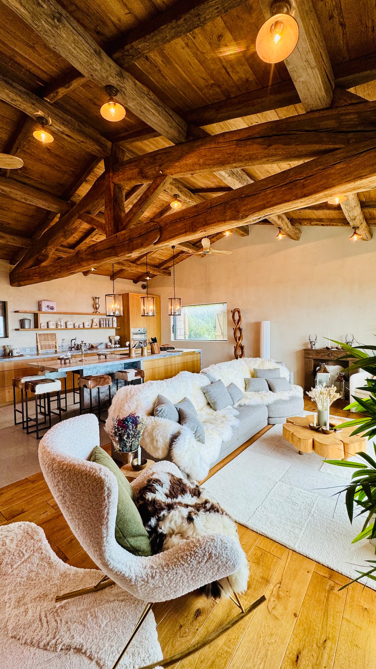 The living area features exposed wooden beams and warm lighting. A plush seating arrangement invites relaxation, complemented by a cozy area rug. A wooden coffee table sits at the center, while the open kitchen is visible in the background, showcasing modern appliances and a natural aesthetic.