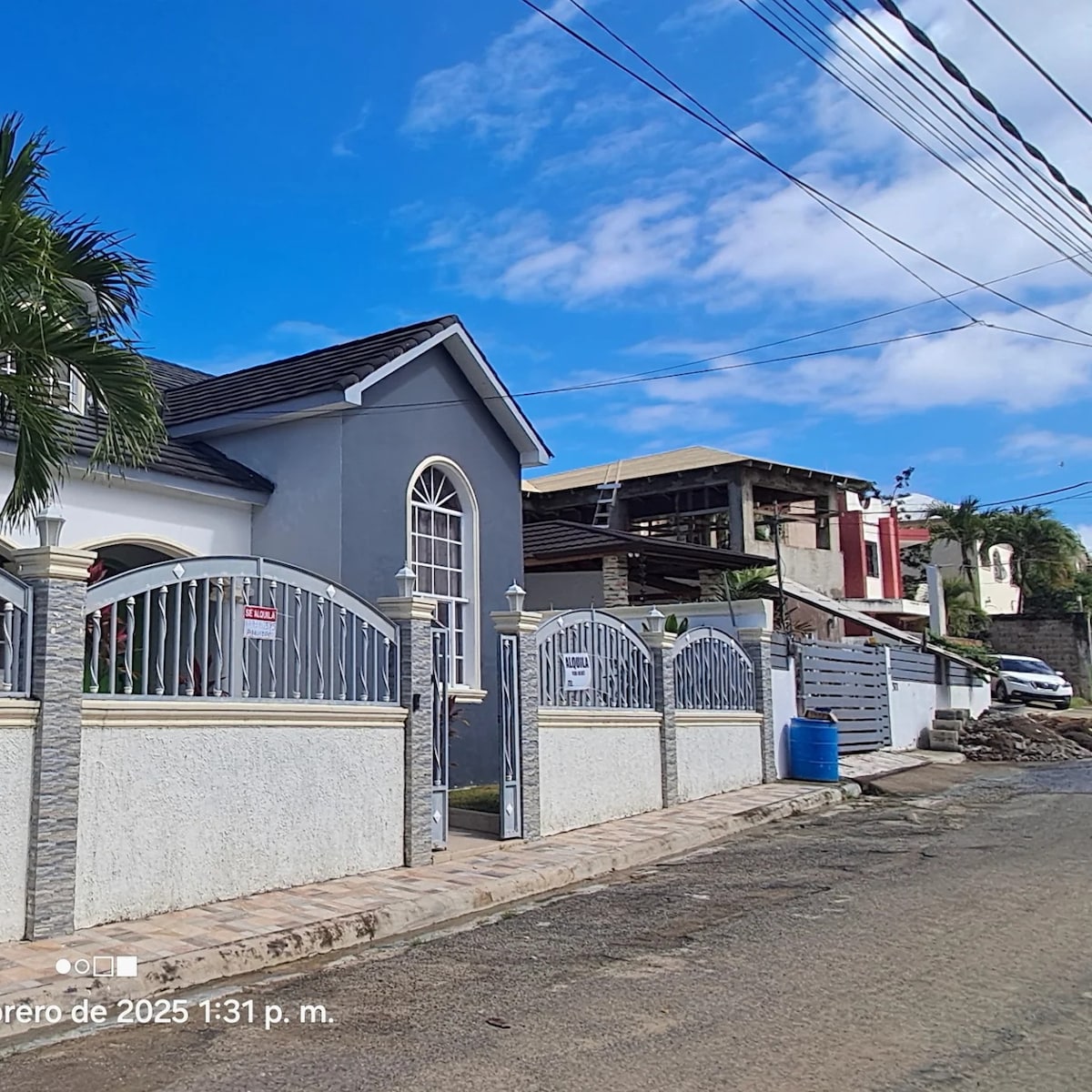 A modern gray house is featured, set against a bright blue sky with scattered clouds. The property is enclosed by an elegant gate, flanked by palm trees, and a small driveway is visible. Nearby structures include a mix of residential buildings, creating a welcoming neighborhood ambiance.