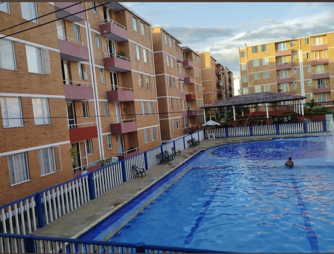 A large swimming pool is set in the foreground, bordered by a decorative fence. Multiple residential buildings with balconies rise in the background, featuring a warm brick facade. A person can be seen enjoying the pool, indicating a welcoming community space.