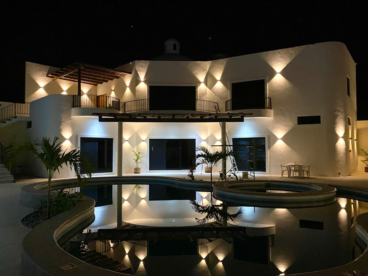 The villa features a striking architectural design, highlighted by strategic exterior lighting. Reflections dance on the calm water of the adjacent pool, which is surrounded by modern landscaping. Expansive windows provide a glimpse of the interior, enhancing the serene evening ambiance.