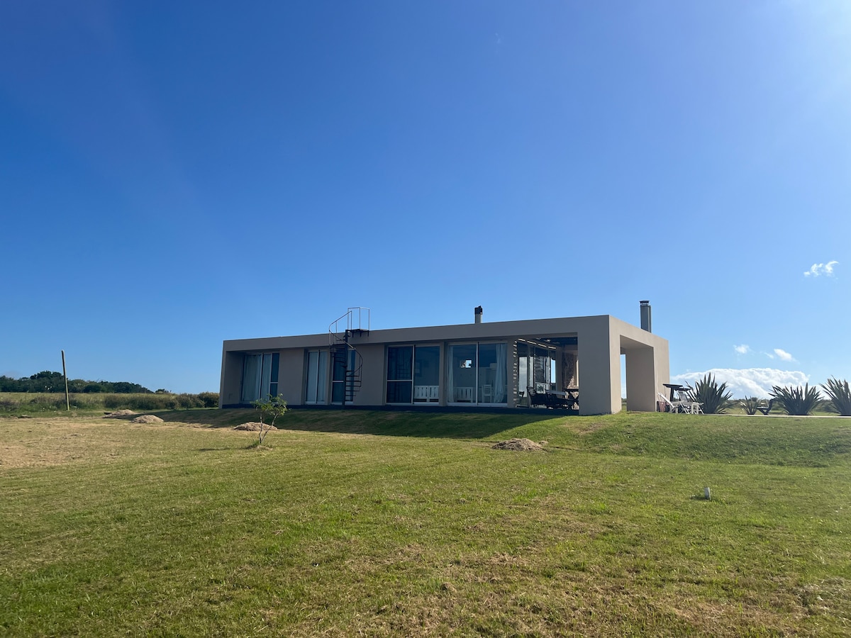 A modern single-story house is set on a grassy landscape, featuring large glass windows and a spiral staircase leading to a rooftop area. The structure is complemented by a serene sky and surrounding nature.