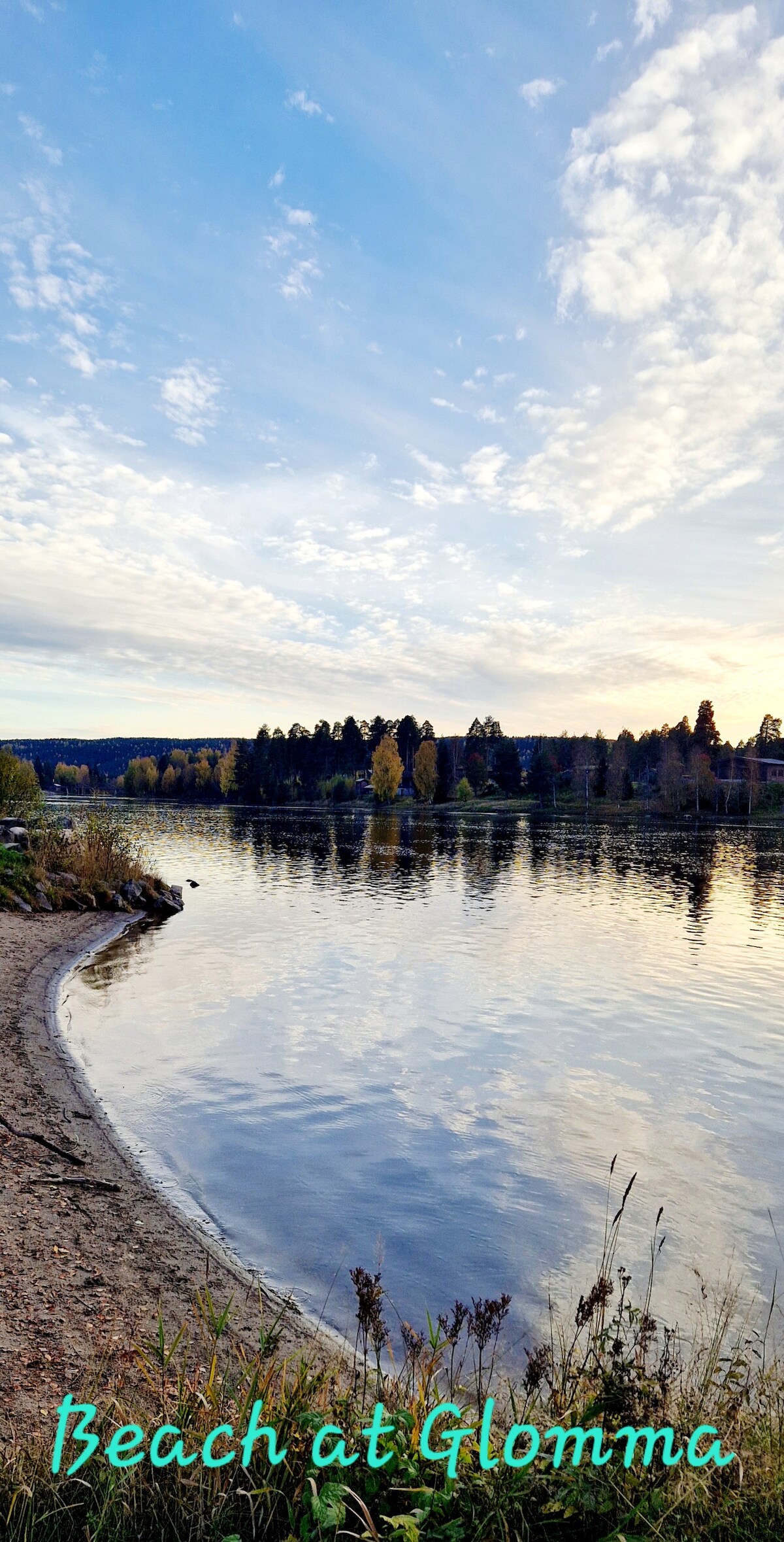 A tranquil beach scene along the Glomma River is depicted, with soft sandy shores meeting calm water. The sky is adorned with scattered clouds, reflecting hues of blue and orange as the sun sets, surrounded by trees in autumn colors.